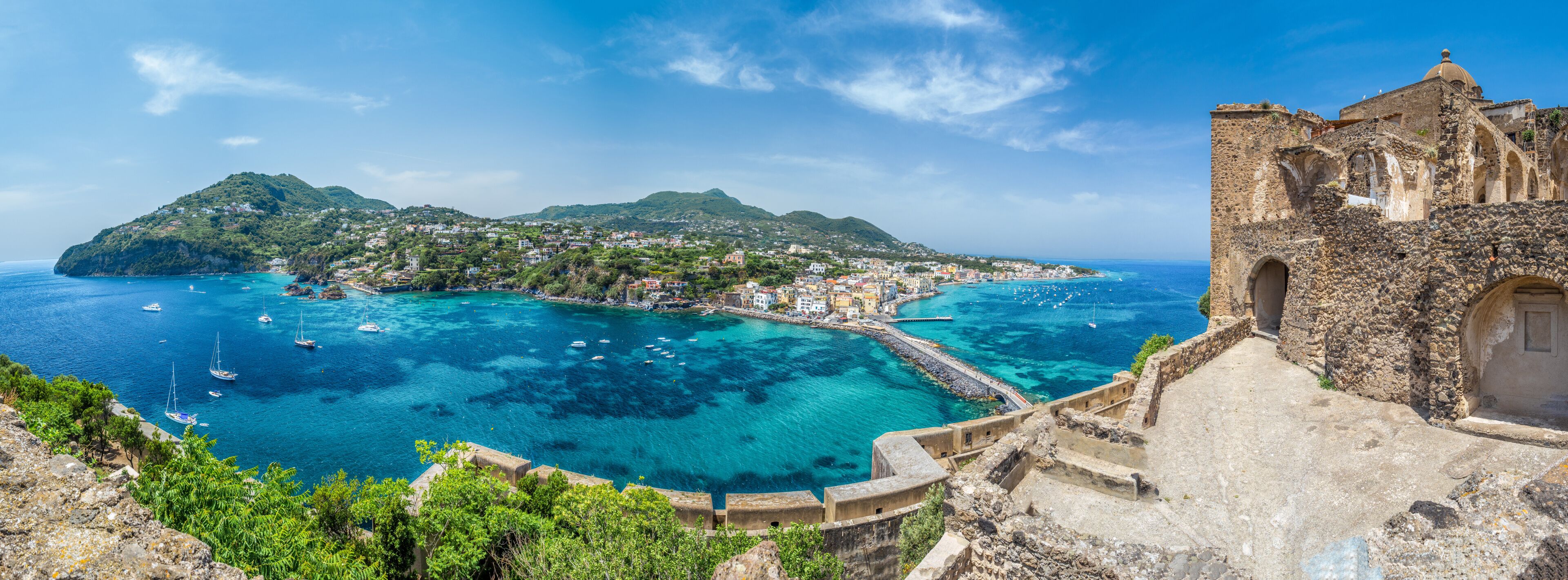 A coastal town with clear blue waters, a historic stone building, and a hilltop with greenery.