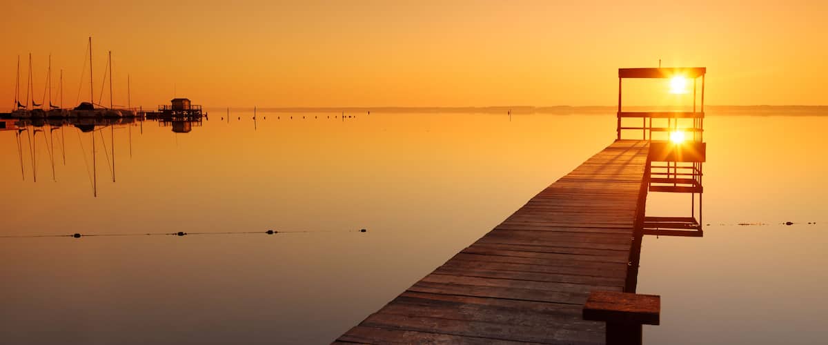 A wooden pier extending into calm waters at sunrise with boats docked on the left and a lighted structure at the end.