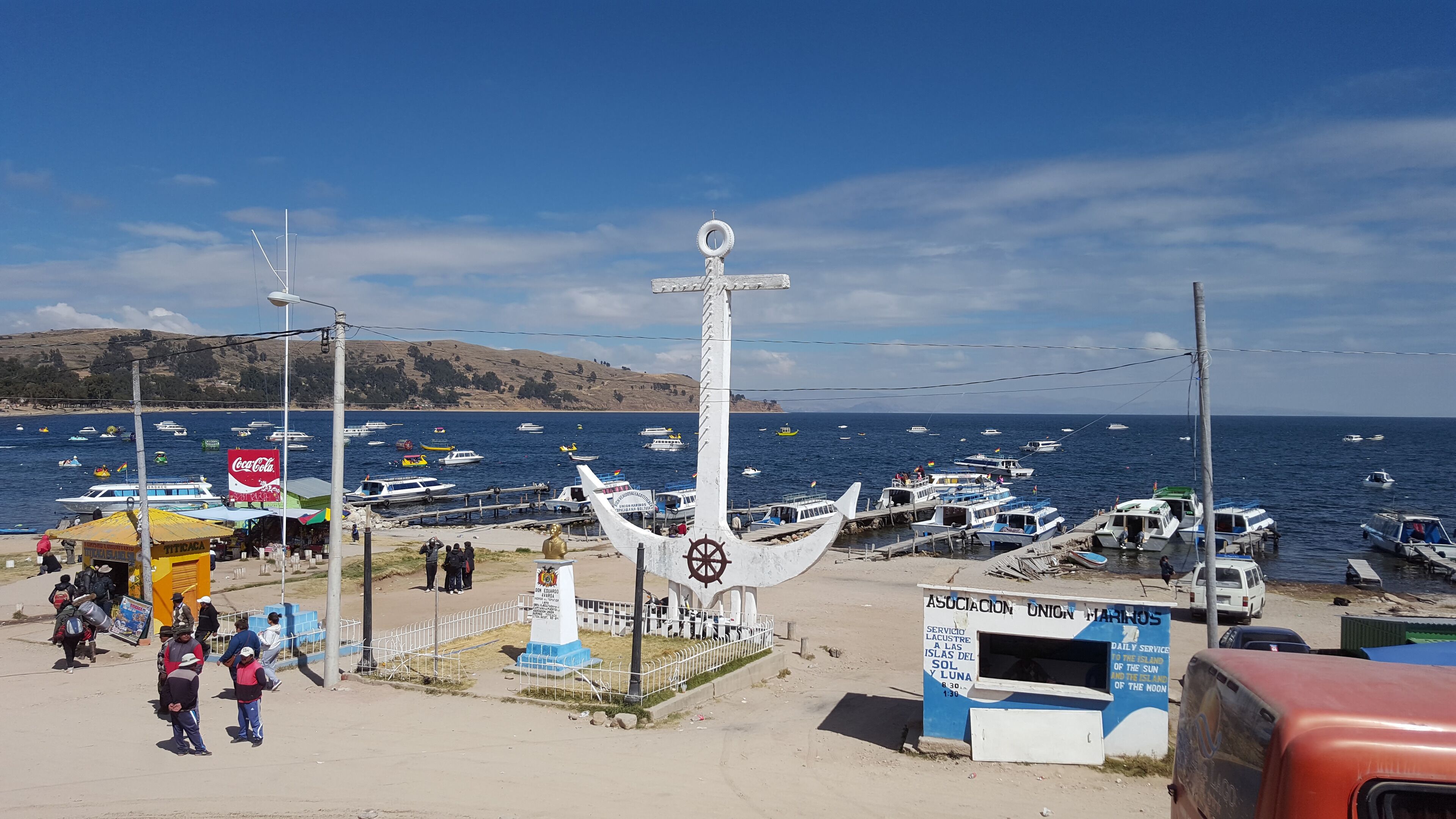 A coastal scene with a prominent white anchor sculpture, a Coca-Cola sign, and several boats docked at a pier.
