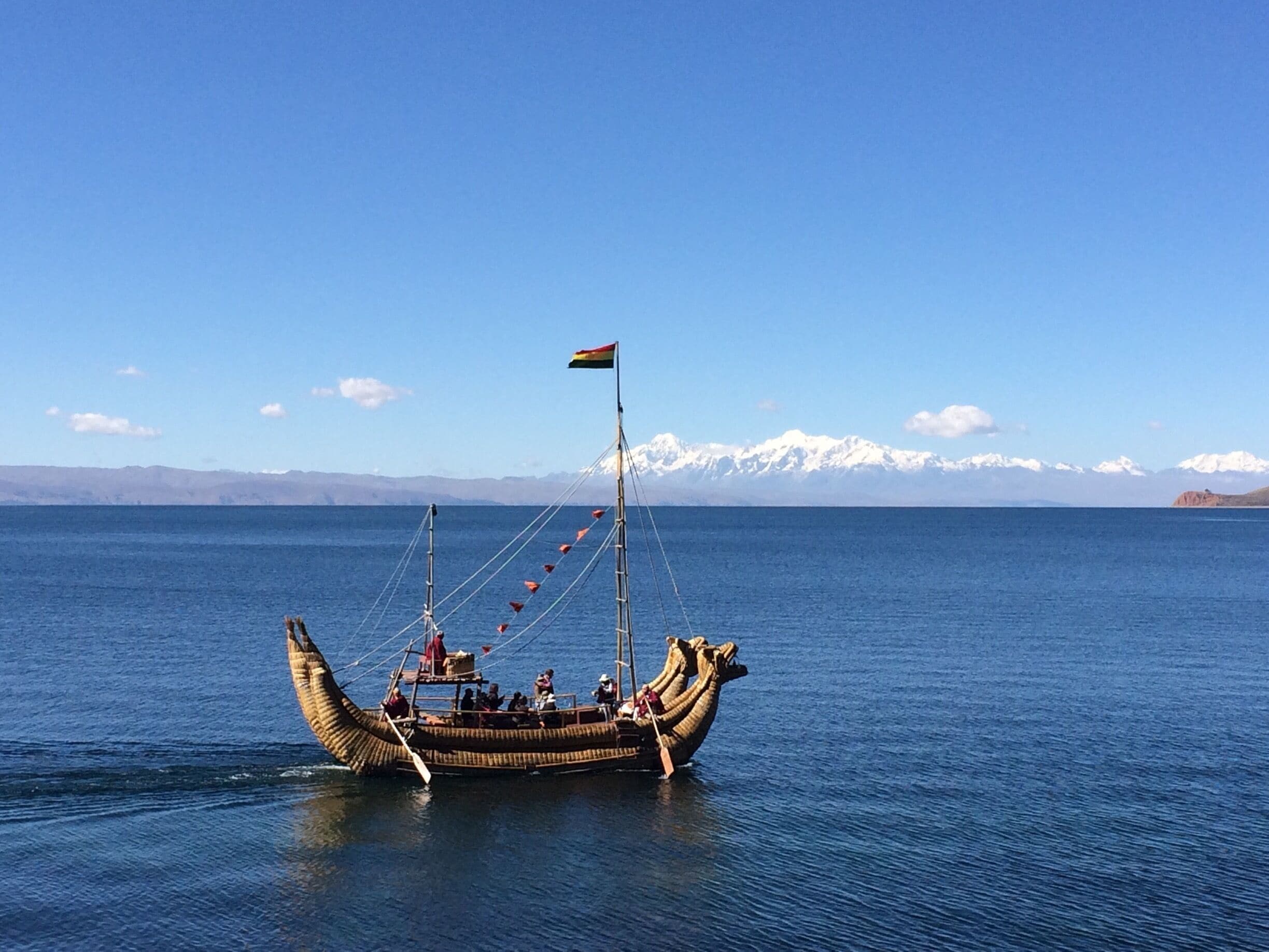 A traditional boat with a flag on a calm sea with snow-capped mountains in the background.