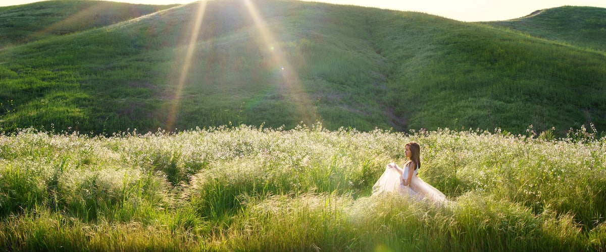 A person in a white dress sitting in a field of wildflowers with hills in the background.