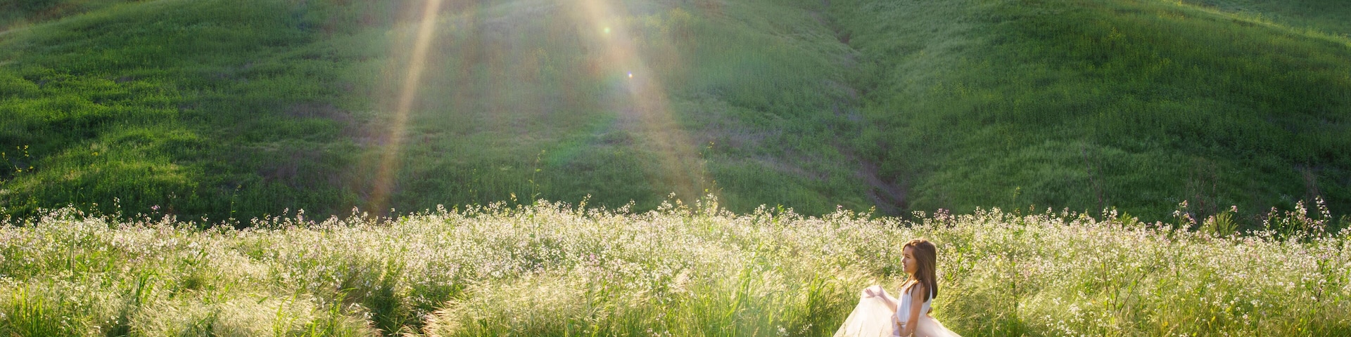 A person in a white dress sitting in a field of wildflowers with hills in the background.