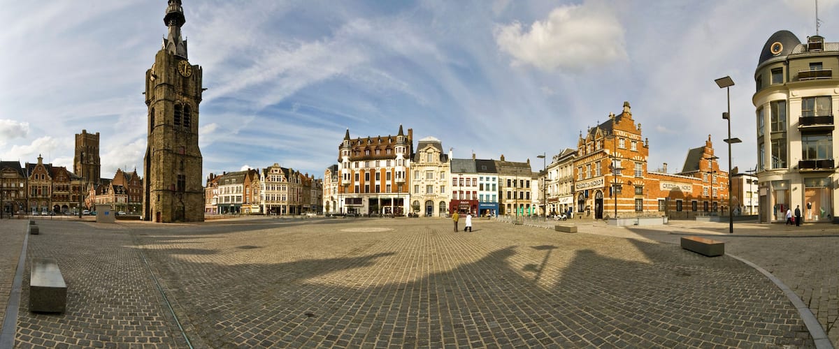 A cobblestone square with historic buildings, a prominent clock tower, and a clear sky.