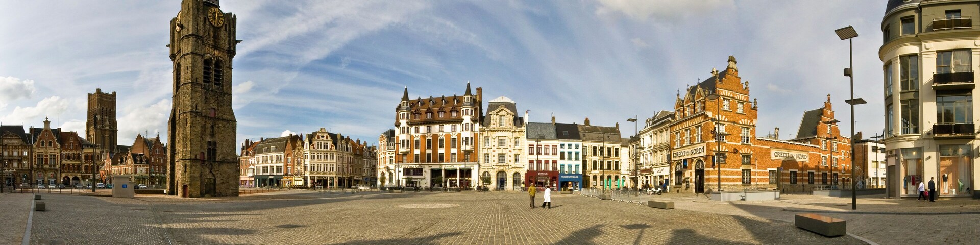 A cobblestone square with historic buildings, a prominent clock tower, and a clear sky.