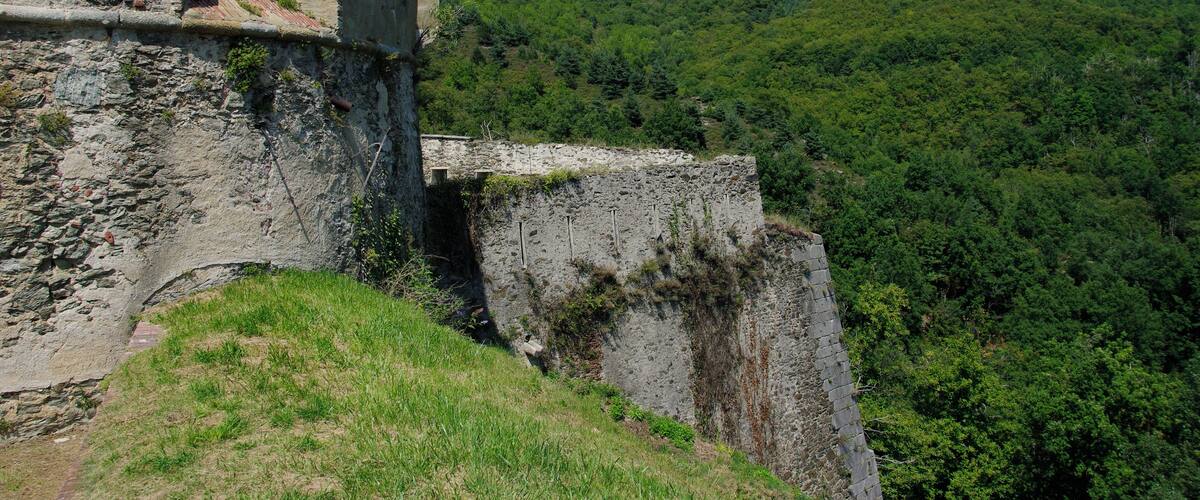 A historic stone fortress with arched windows and a steep cliff overlooking a dense forest.