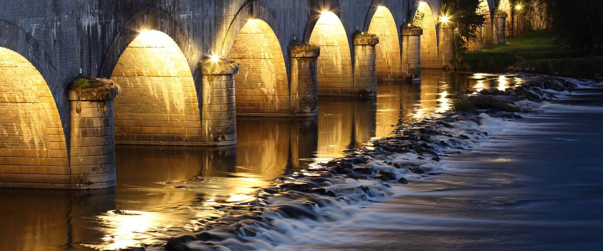 A stone bridge with illuminated arches over a flowing river at dusk.