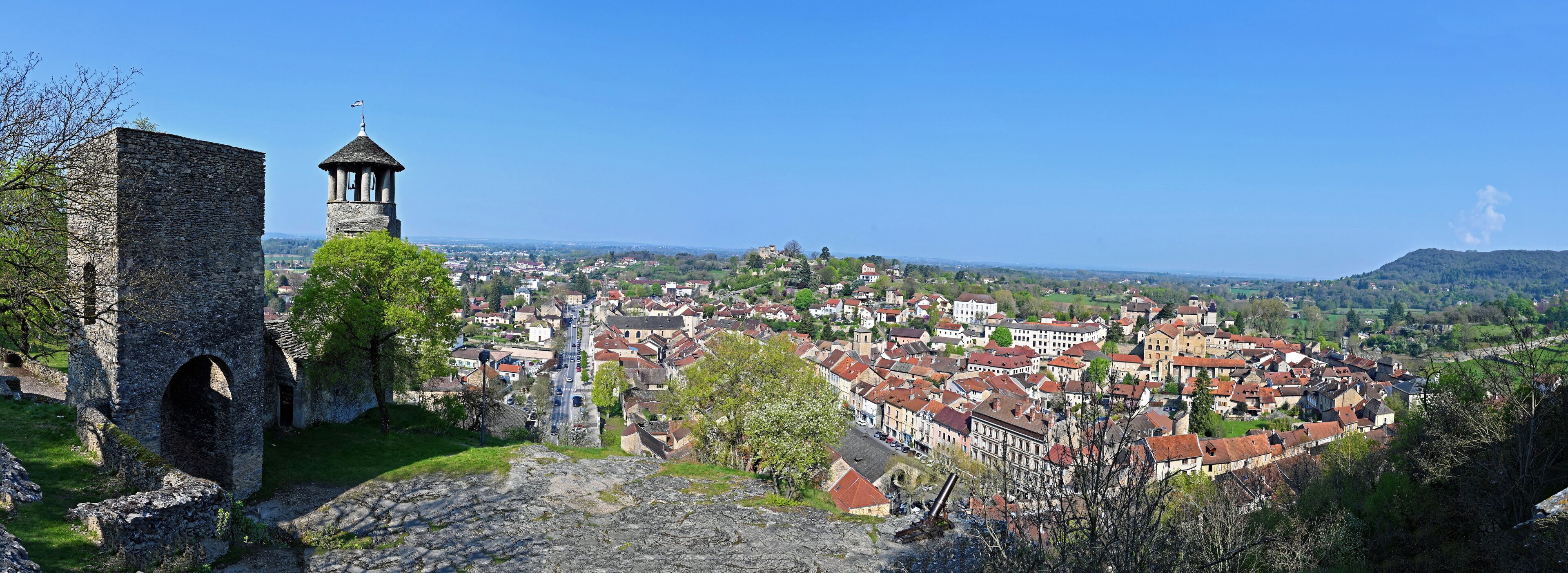 A historic stone tower with a weather vane, a cobblestone path, and a view of a town with red-roofed buildings.