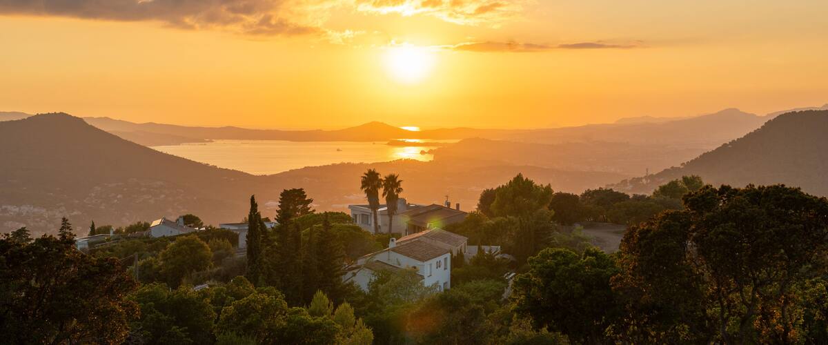 A sunset over a coastal landscape with mountains, a body of water, and residential buildings.