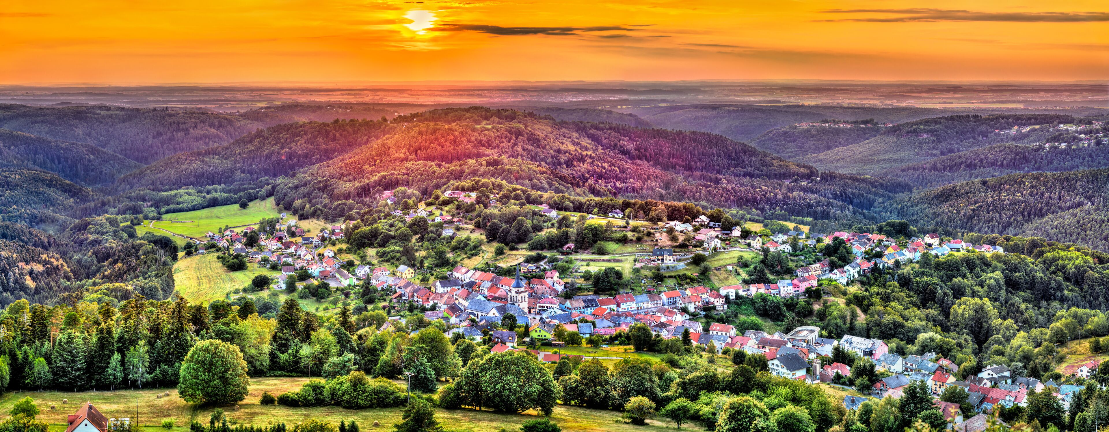 A village nestled in a valley with rolling hills and a sunset in the background.