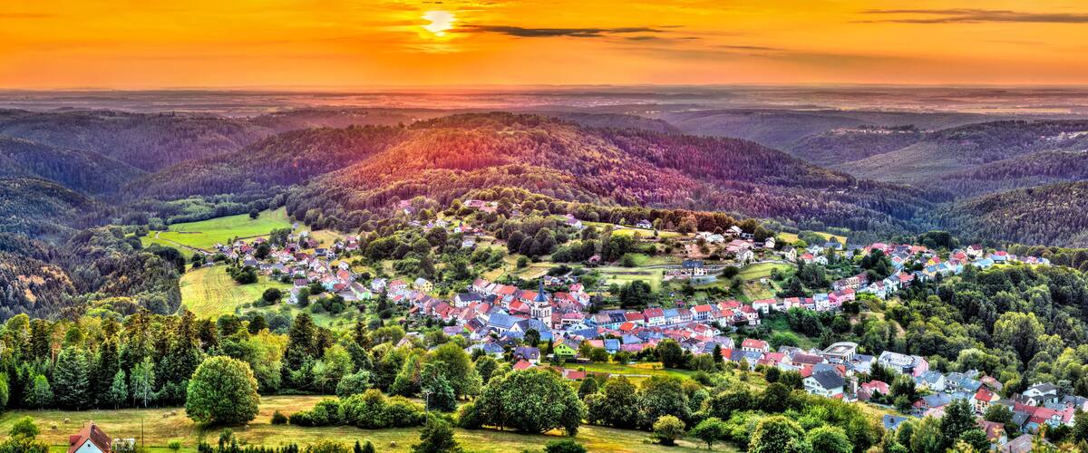 A village nestled in a valley with rolling hills and a sunset in the background.