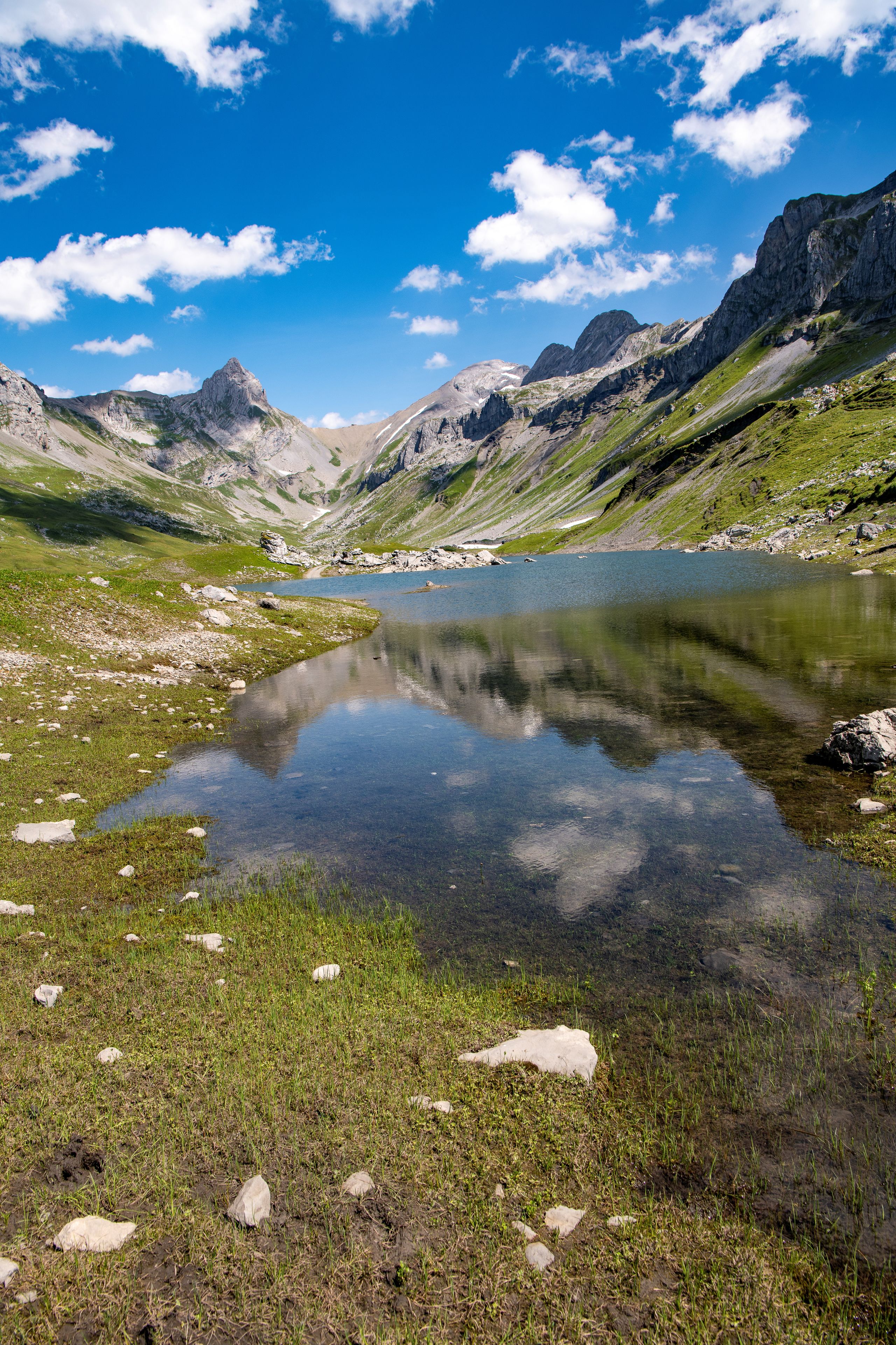 Un lago de montaña con reflejos cristalinos, rodeado de terreno rocoso y vegetación verde.
