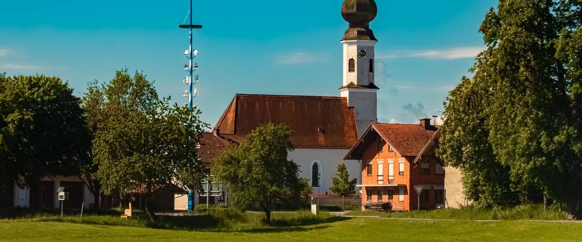 A church with a tall bell tower and a weather vane, surrounded by trees and a clear blue sky.