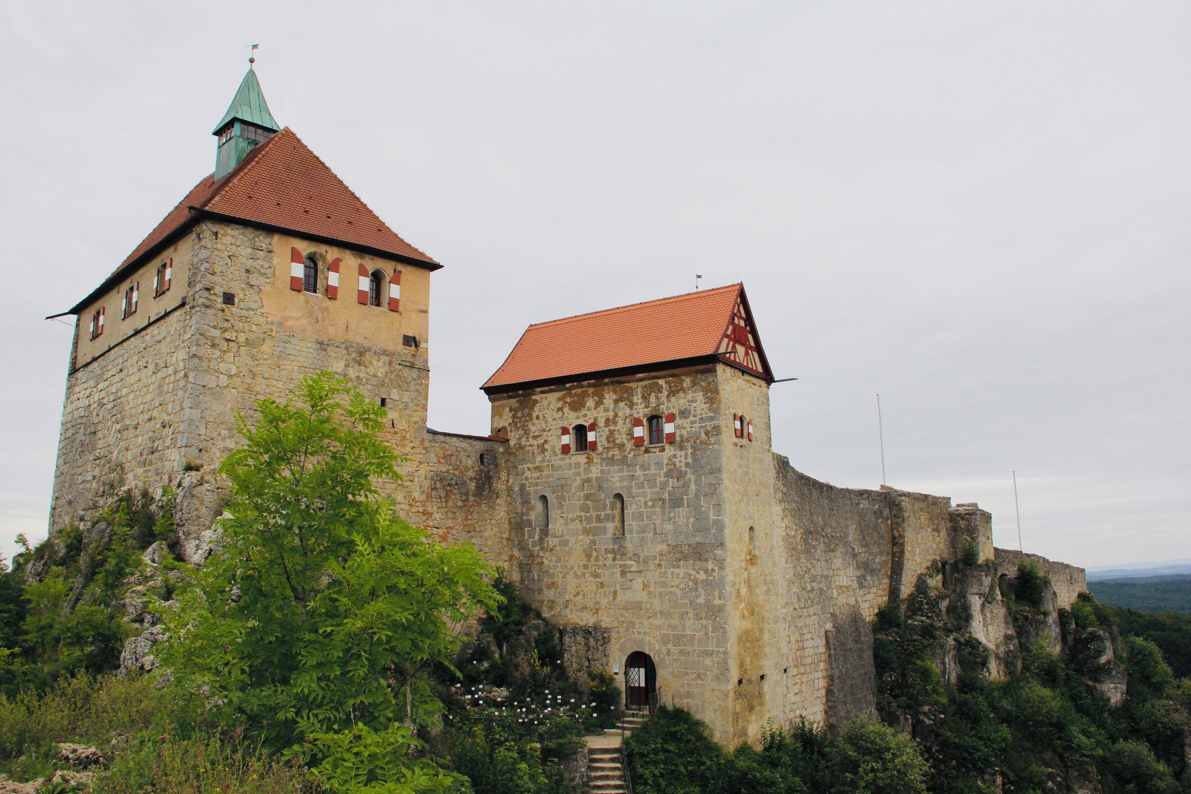 A historic stone castle with a red-tiled roof, a tower with a green spire, and a staircase leading to the entrance.