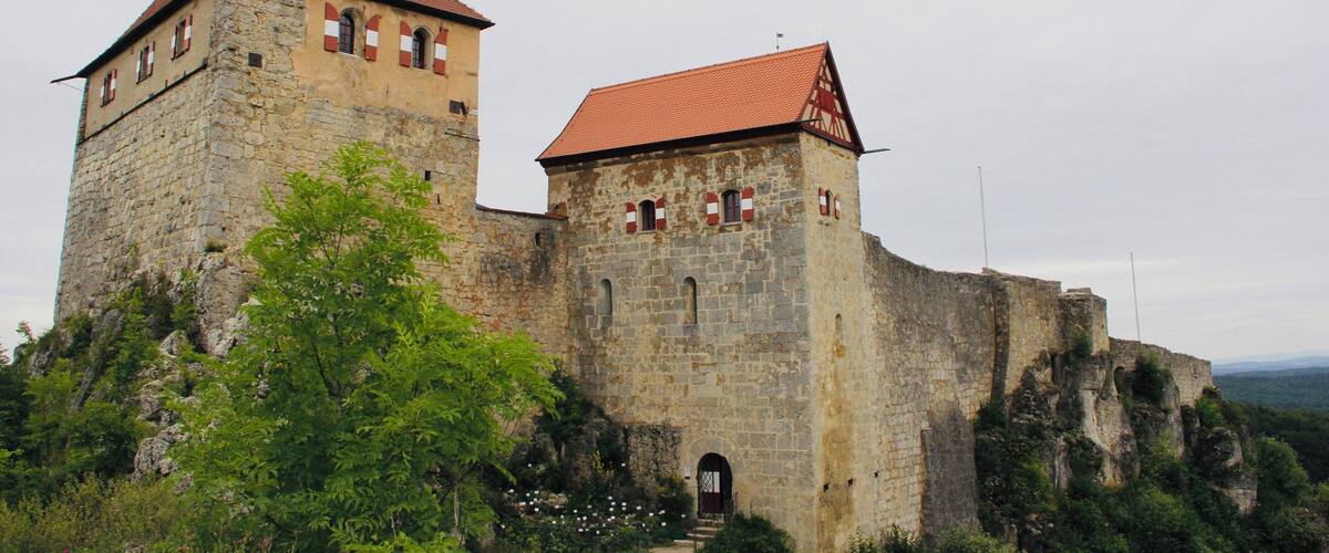 A historic stone castle with a red-tiled roof, a tower with a green spire, and a staircase leading to the entrance.
