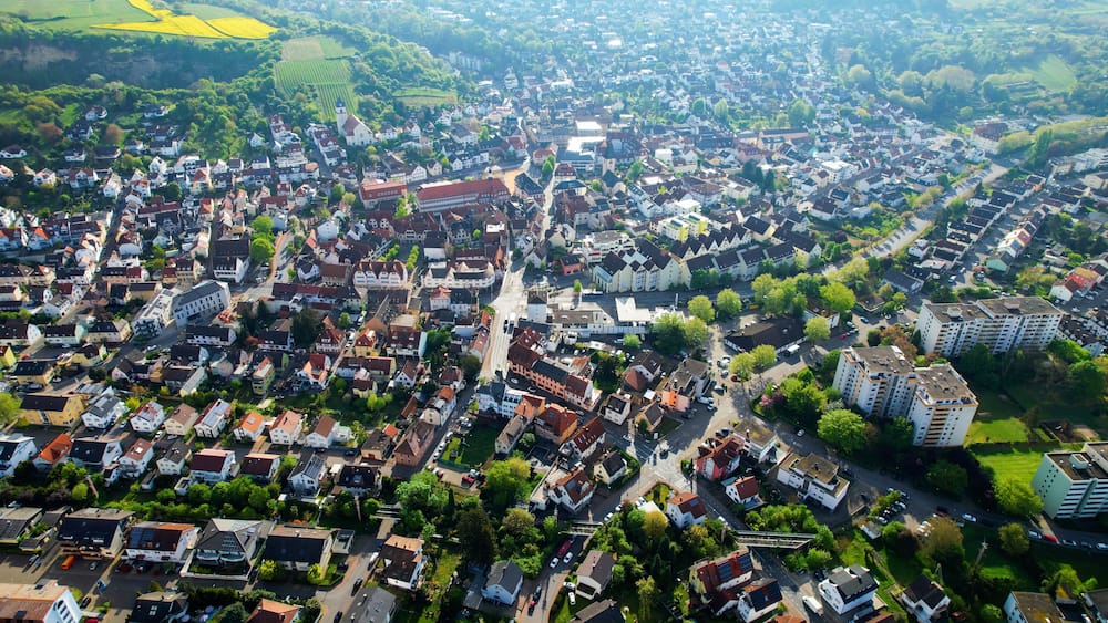 An aerial view of a densely populated town with a mix of residential houses and modern buildings, surrounded by green fields and a prominent church tower.