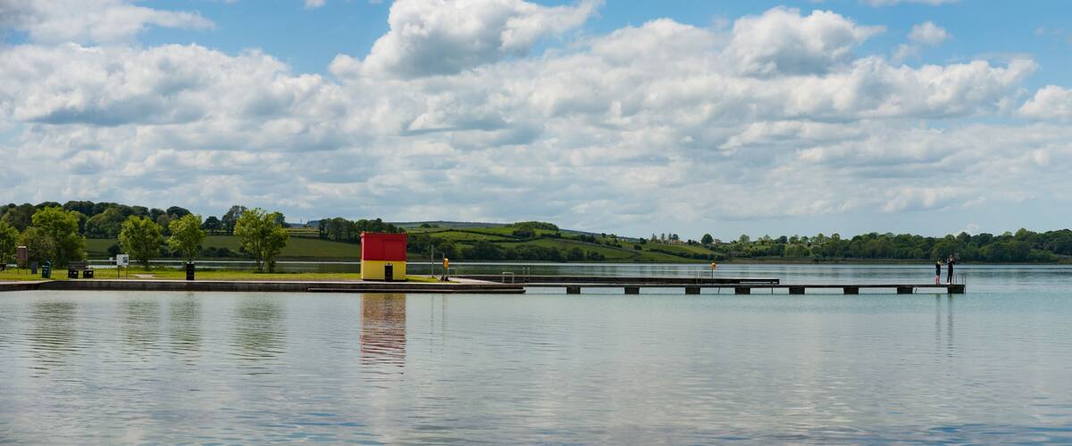 A calm lake with a dock extending into the water, surrounded by green hills and a partly cloudy sky.
