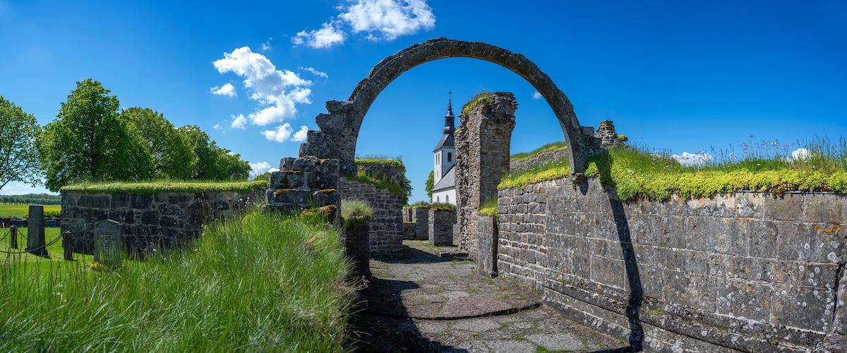 An ancient stone archway leading to a church with a cross on top, surrounded by lush greenery and a clear blue sky.