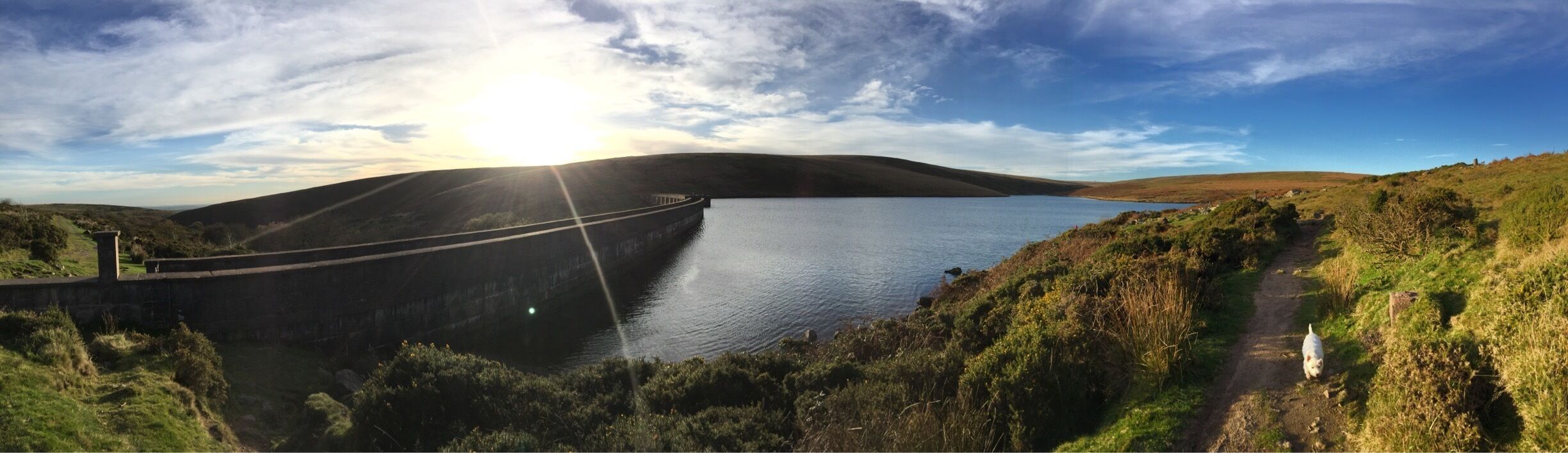 A dam with a reservoir, surrounded by hills and a clear sky.