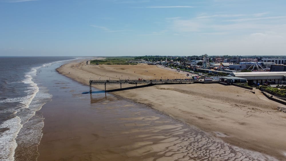 A beach with a pier extending into the ocean, a sandy shore, and a distant town with buildings and a Ferris wheel.