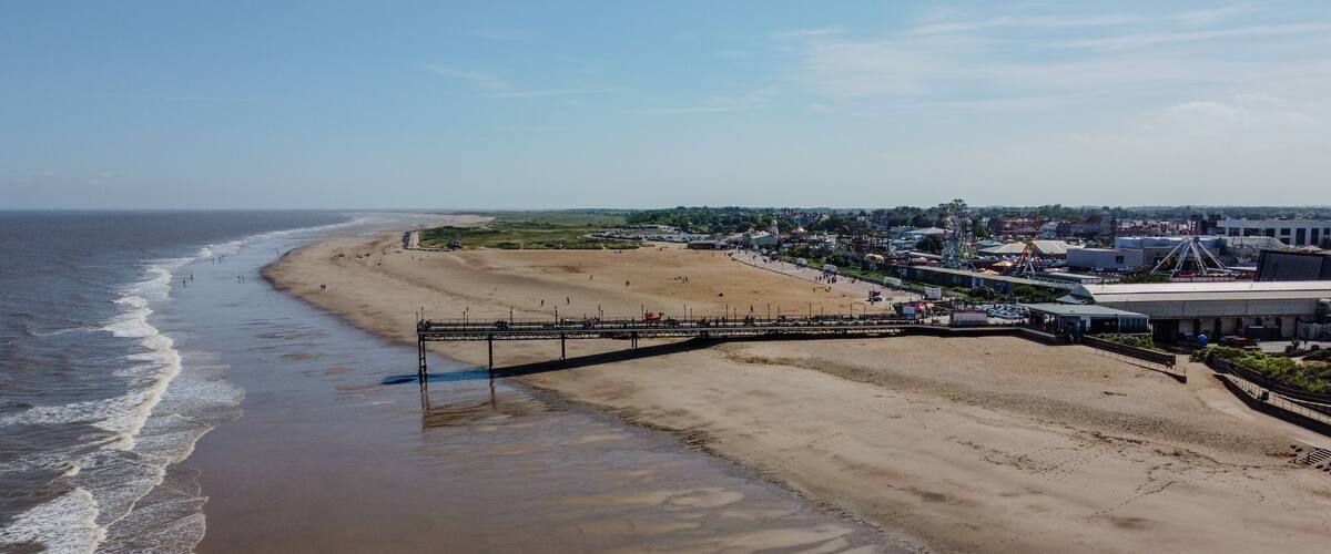 A beach with a pier extending into the ocean, a sandy shore, and a distant town with buildings and a Ferris wheel.