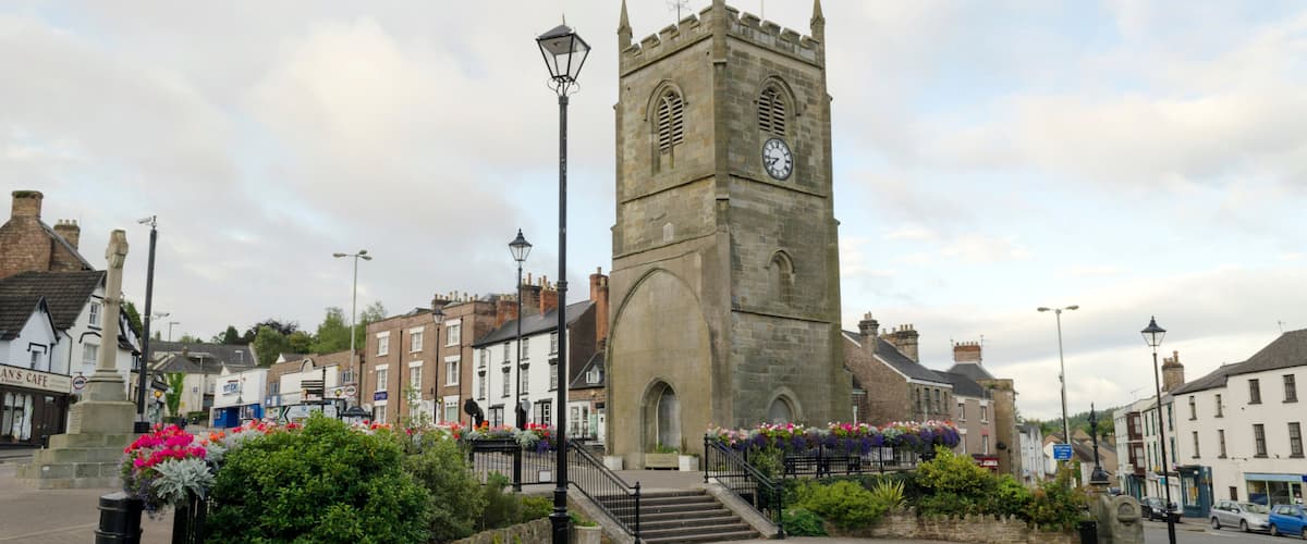 A historic tower with a clock, surrounded by cobblestone streets and flower beds.