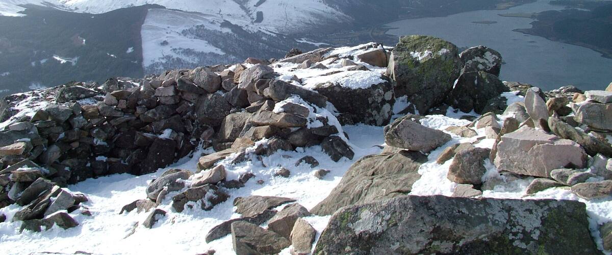 Paisaje nevado de montañas con afloramientos rocosos y un lago a lo lejos.