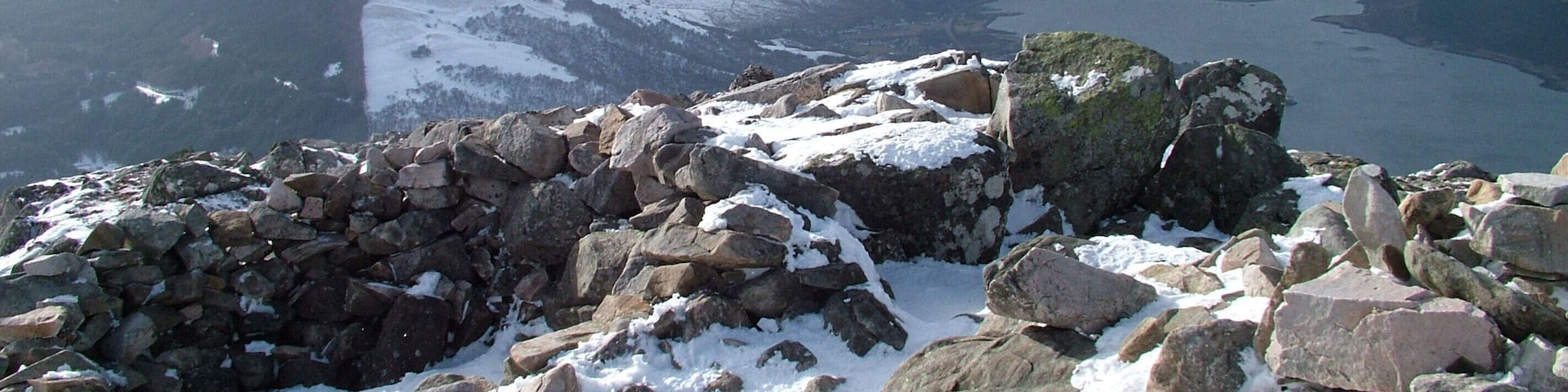 A snowy mountain landscape with rocky outcrops and a lake in the distance.