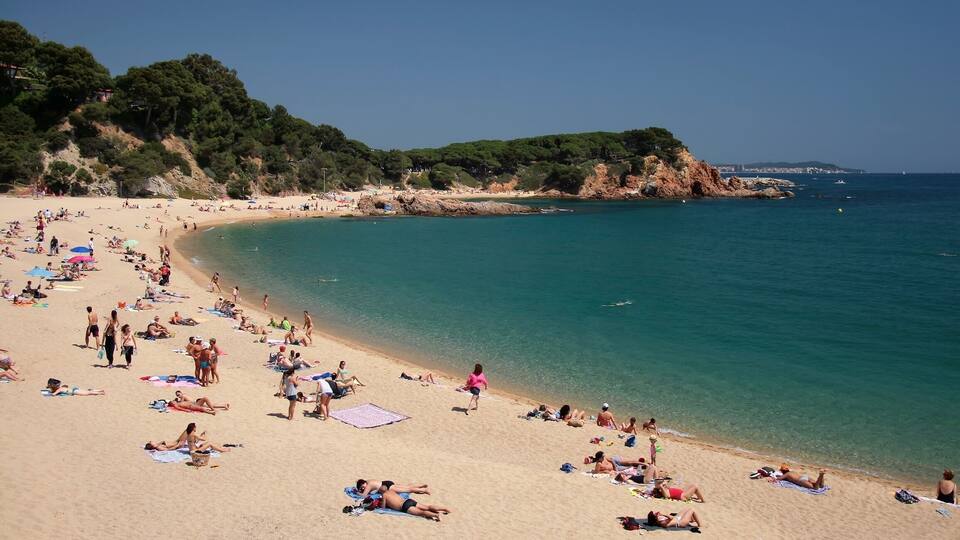 A crowded beach with people sunbathing and swimming, surrounded by rocky cliffs and clear blue water.