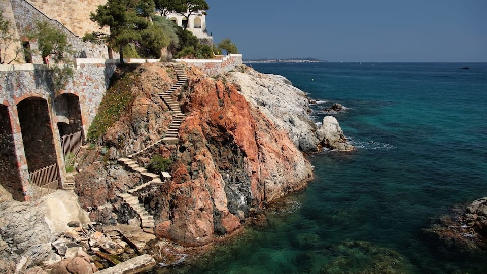 A coastal stone building with a staircase leading down to the sea, clear turquoise water, and a distant city skyline.