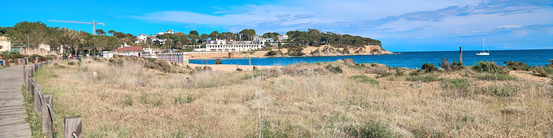 Un paisaje costero con un paseo marítimo de madera, una playa arenosa, un mar azul cristalino y una colina con edificios y árboles.