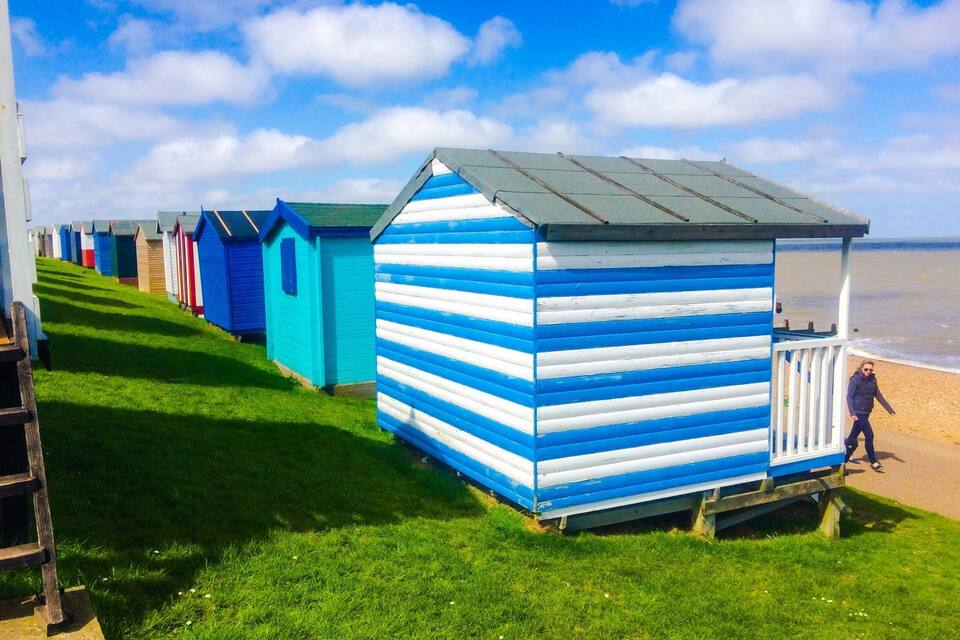 A row of colorful beach huts along a grassy promenade by the sea.