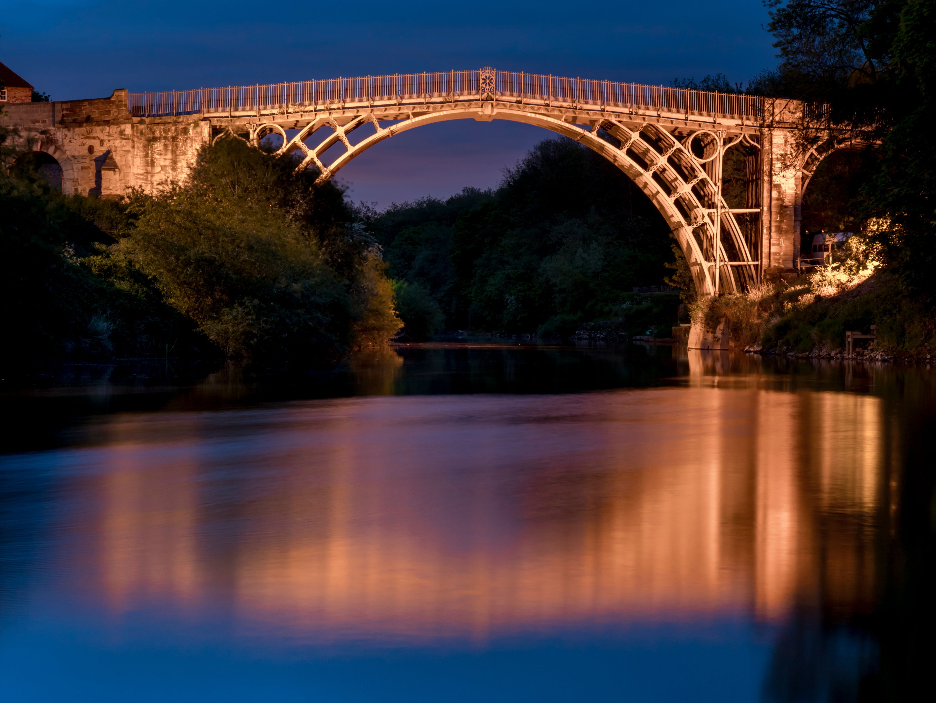 Eine steinerne Brücke mit Metallgeländer, die über einen ruhigen Fluss führt, bei Nacht.