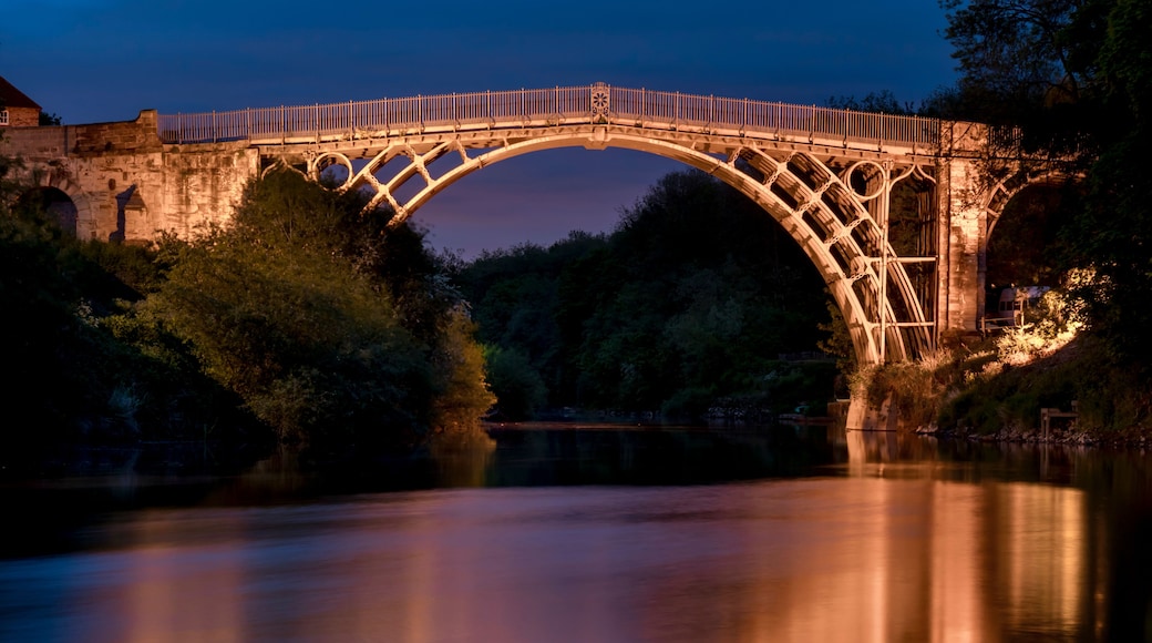 Un puente de piedra con barandilla metálica, arqueándose sobre un río tranquilo por la noche.