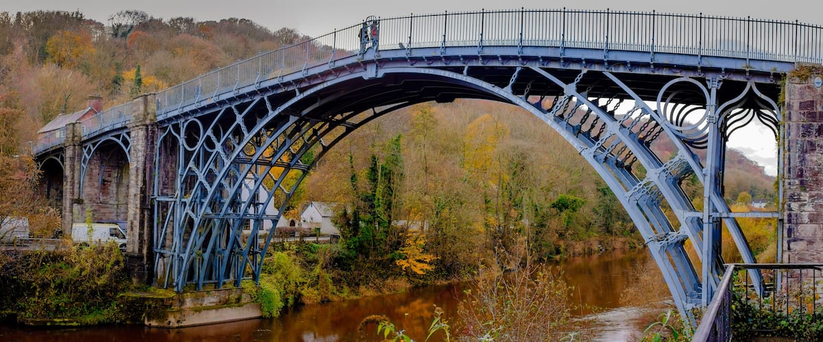 Un puente metálico con arco sobre un río, rodeado de follaje otoñal y edificios de piedra.