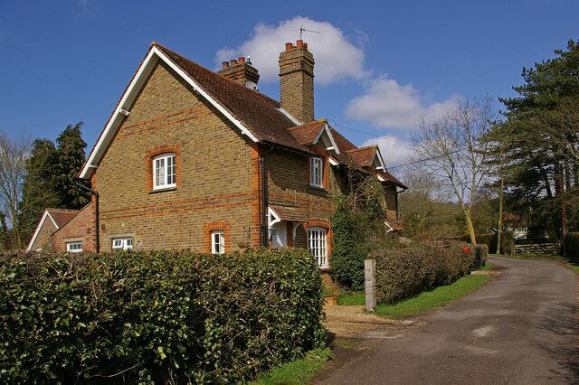 A traditional brick house with a sloped roof and a chimney, surrounded by a hedge and a narrow road.
