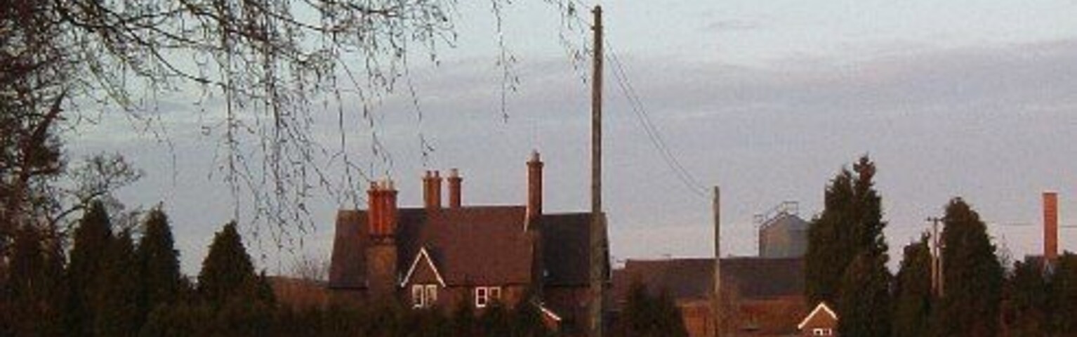 Paisaje rural con un campo extenso, una casa y chimeneas industriales al fondo.