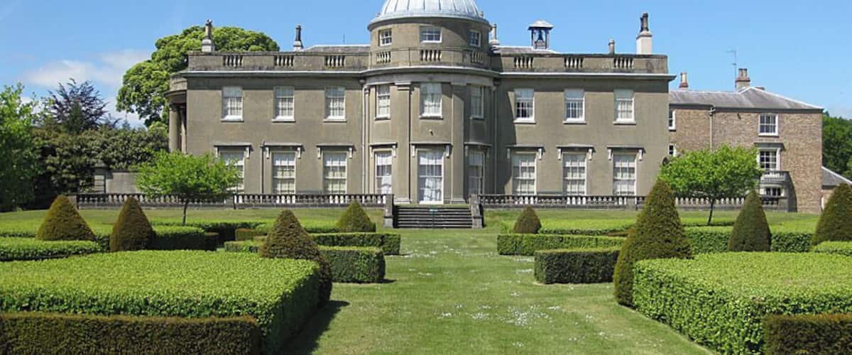 Un edificio grande y señorial con una cúpula central, rodeado de setos cuidados y un jardín formal.