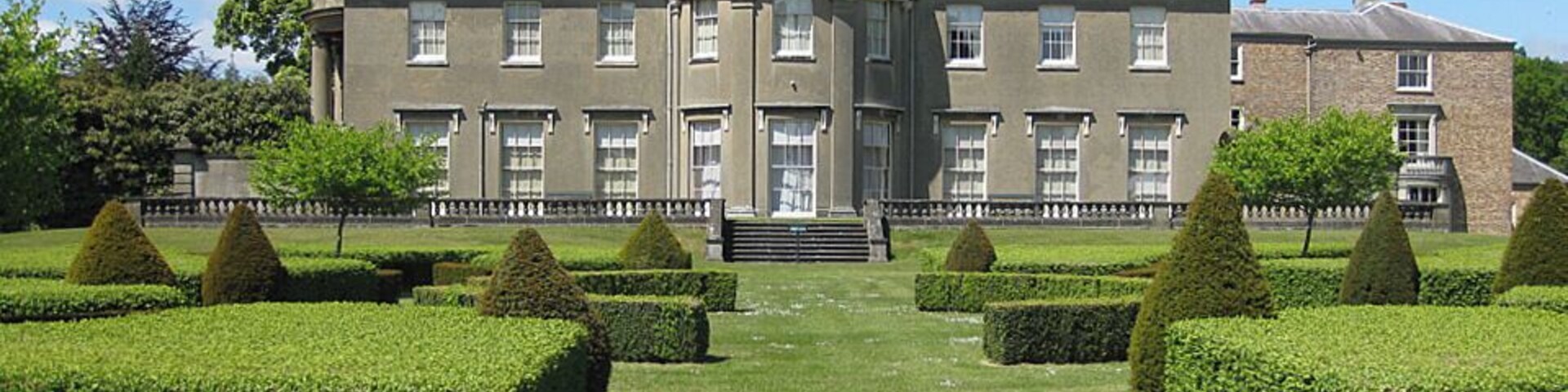 Un edificio grande y señorial con una cúpula central, rodeado de setos cuidados y un jardín formal.