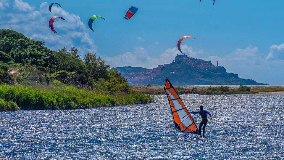 A person windsurfing on a calm body of water with multiple kites in the background.