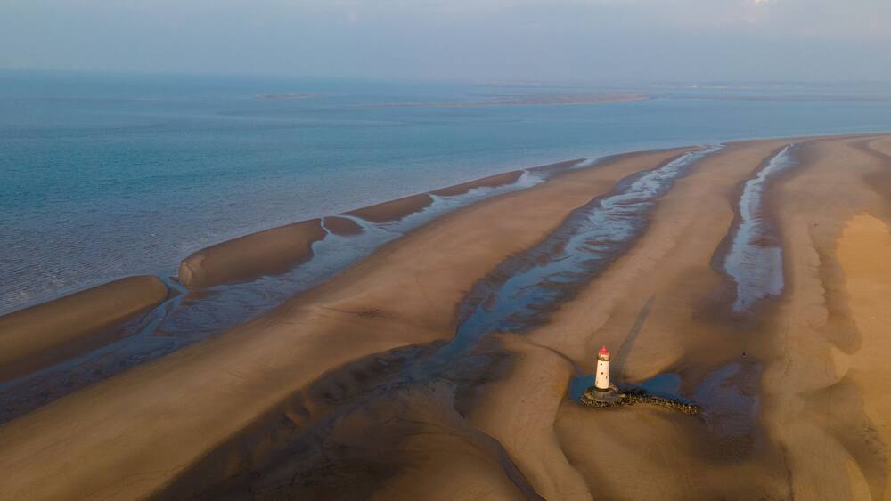 A coastal landscape with sand dunes, a lighthouse, and a calm sea.