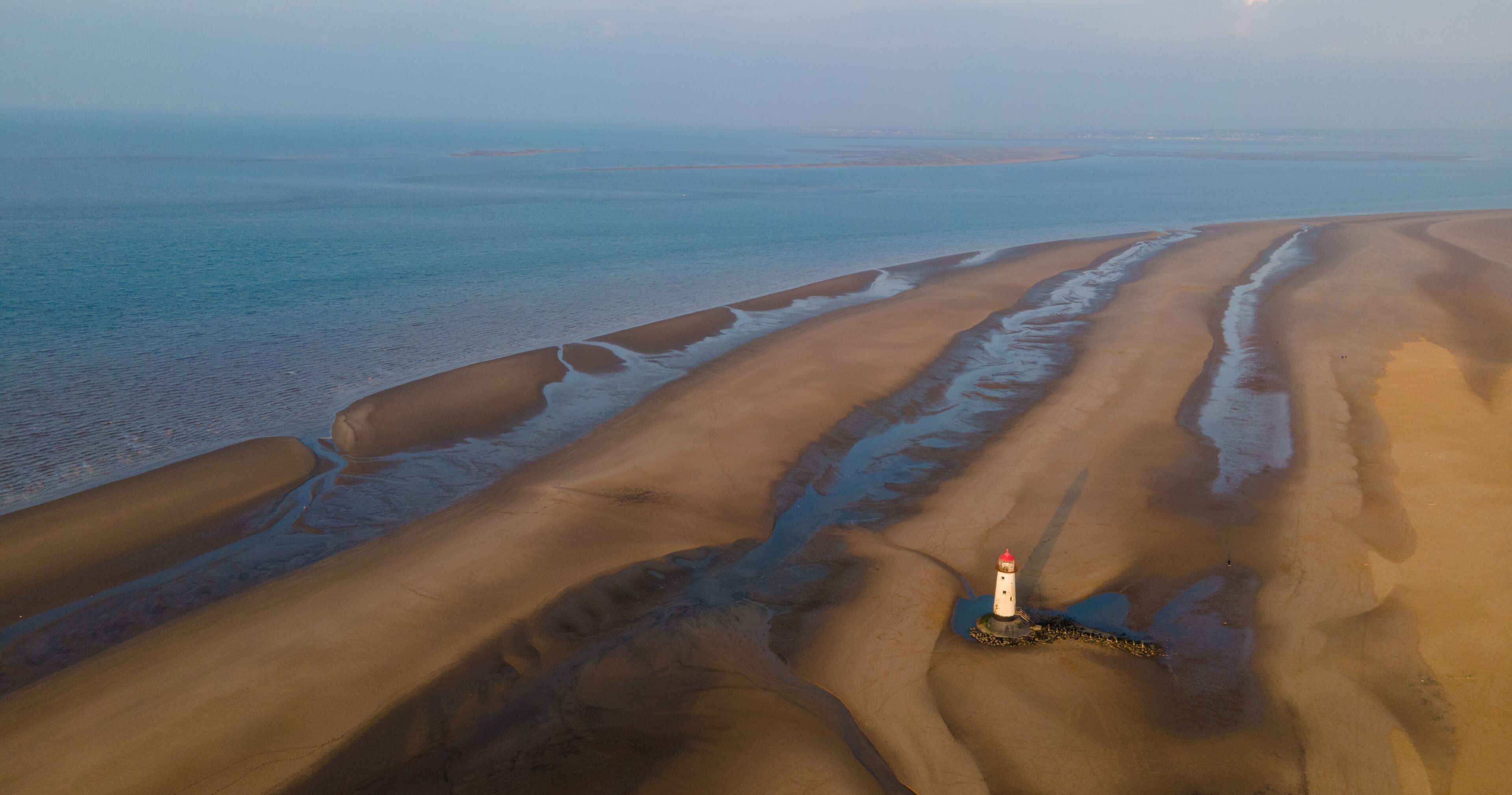 A coastal landscape with sand dunes, a lighthouse, and a calm sea.