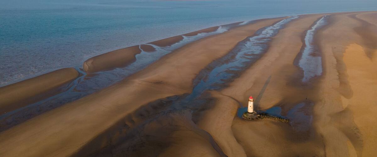A coastal landscape with sand dunes, a lighthouse, and a calm sea.