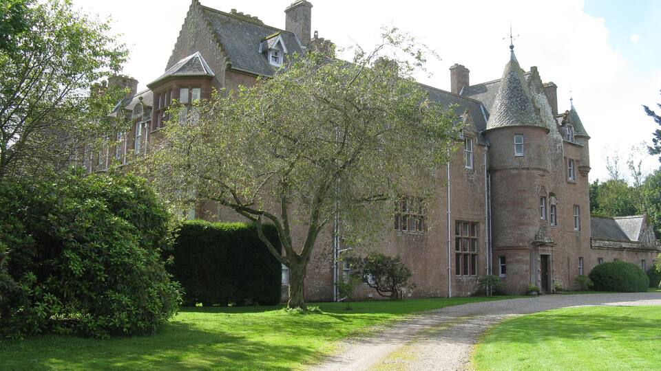 A large, historic stone building with a prominent turret and a well-maintained lawn.
