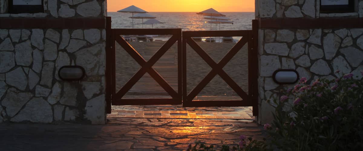 A stone gate with a wooden crossbar, leading to a beach with umbrellas and the sun setting over the ocean.