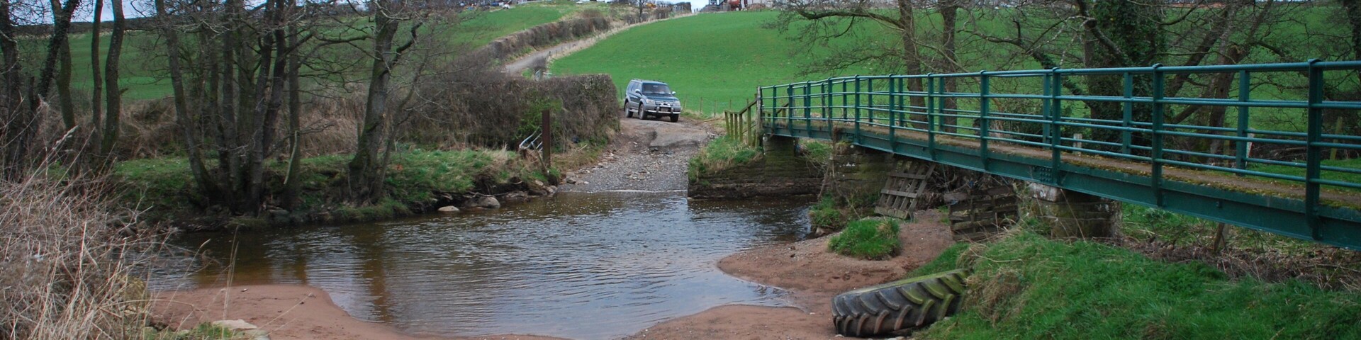Un arroyo rural cruzando un puente estrecho, rodeado de árboles y una colina cubierta de hierba.