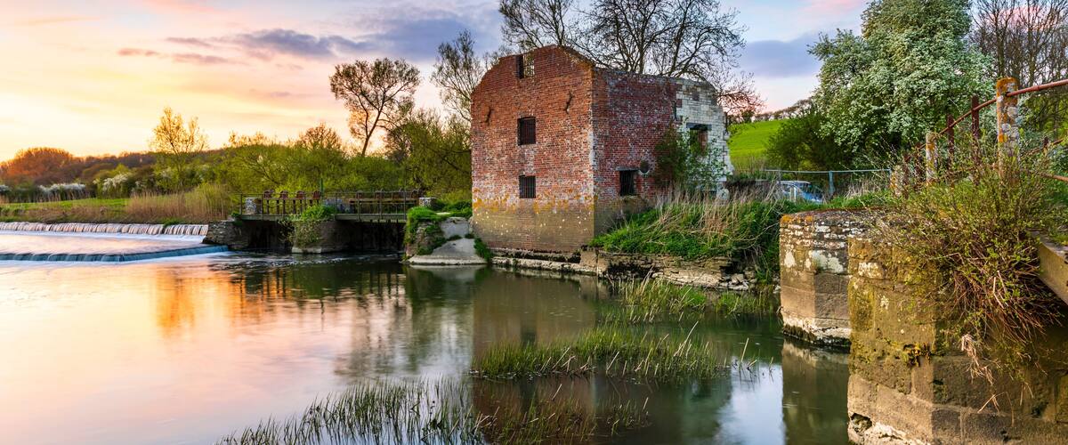 A rustic brick mill by a calm river with a wooden bridge and lush greenery.