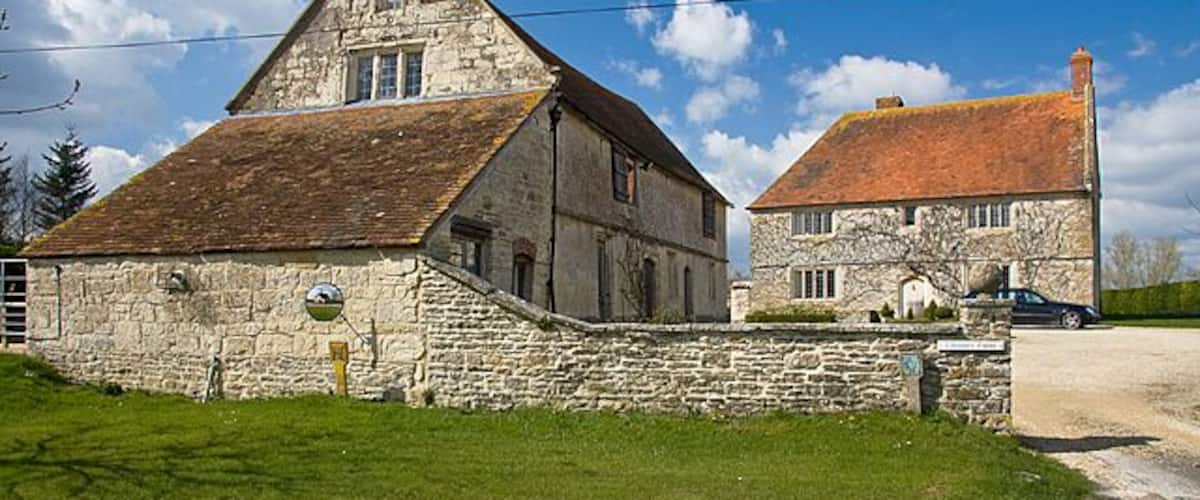 A stone farmhouse with a red-tiled roof, surrounded by a grassy field and a dirt road.