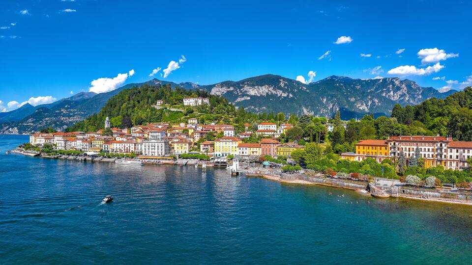 A coastal town with colorful buildings, a calm lake, and mountains in the background.