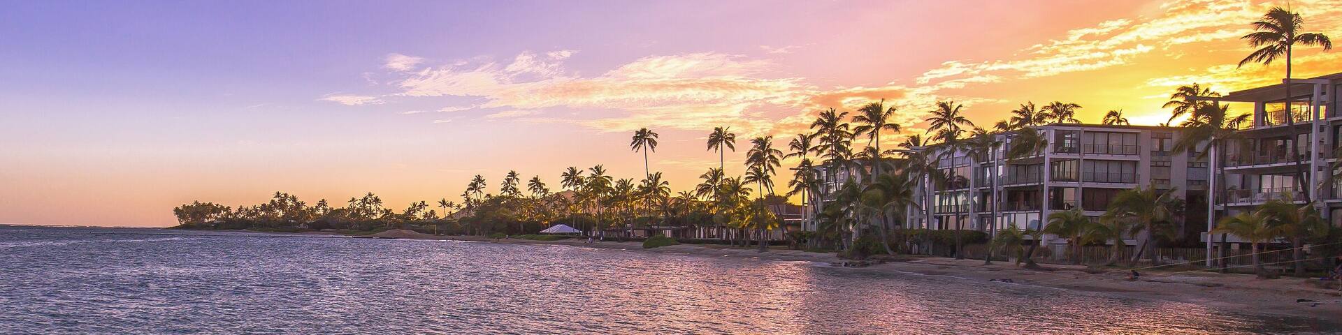 A sunset over a beach with palm trees and a hotel.