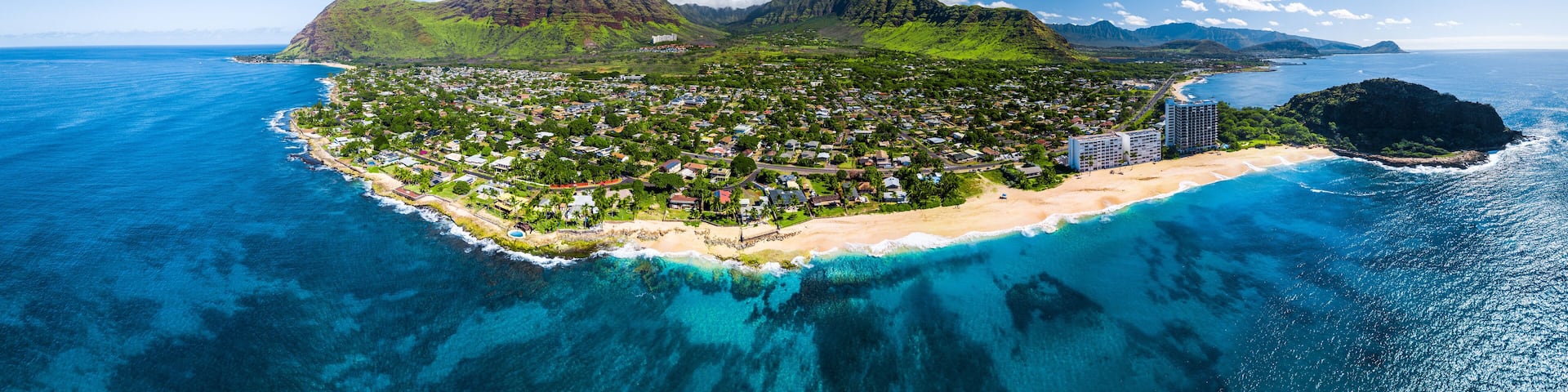 An aerial view of a coastal town with a beach, clear blue waters, and lush green mountains in the background.